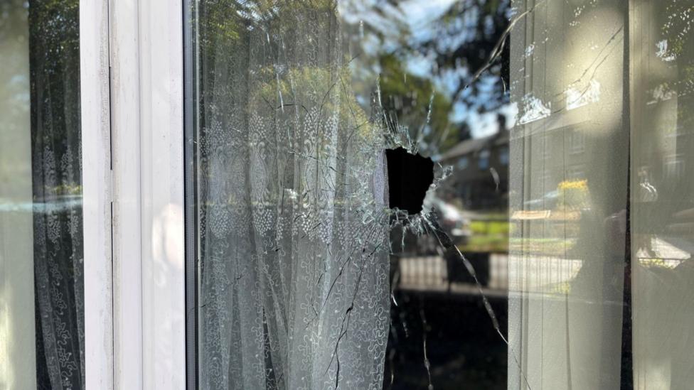 A broken window. There is a large hole in the glass window pane, with cracks radiating out from the centre. Behind the glass a net curtain can be seen hanging down.