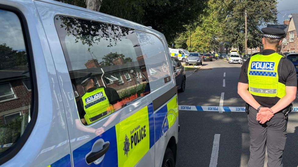 A residential street is cordoned off with bright yellow police tape, with several South Yorkshire Police vehicles parked along the road. A police officer in a high‑visibility vest stands facing the taped‑off area.