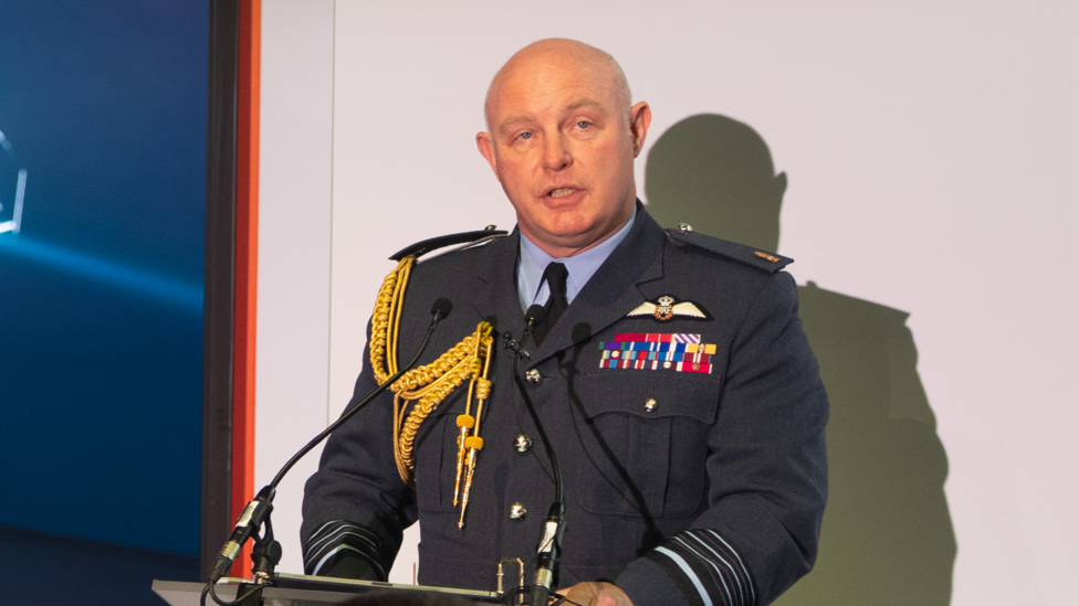 Mid-shot of Air Chief Marshal Sir Harvey Smyth taken in July 2026. He is looking off-camera and speaking behind a podium with a microphone. He is wearing a navy blue military uniform with golden tassels across his shoulder.