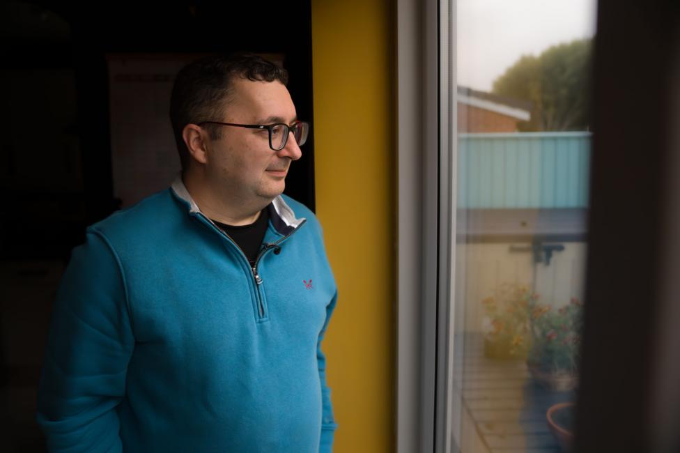 Thornton-Cleveleys resident Andrew Molloy looks out of the window in his home. He is wearing a blue jumper and glasses.