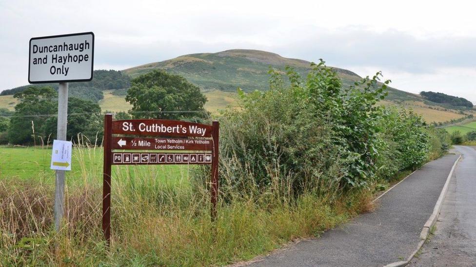 A sign post for the St Cuthbert's Way with a pavement heading off into the hills