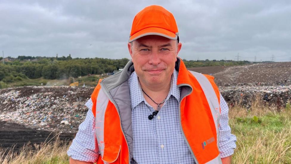 A man in a blue checked shirt and high vis jacket with matching baseball cap standing in front of a landfill site.