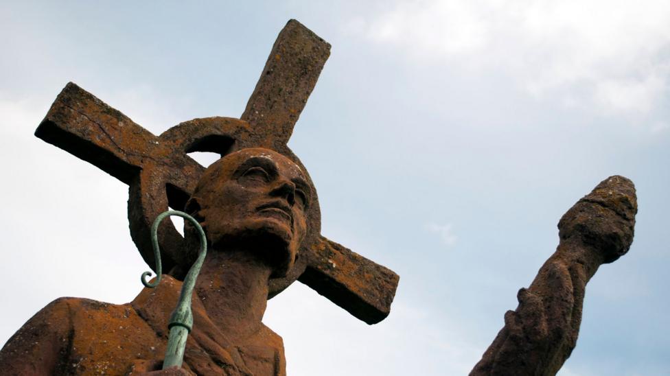 A stone statue of St Cuthbert with a crook in his hand looking off into the distance against a blue sky with a few clouds