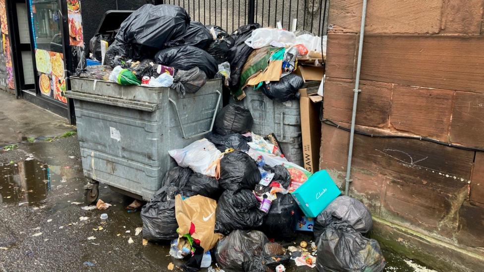 A large grey industrial bin overflowing with rubbish on a city street in Glasgow