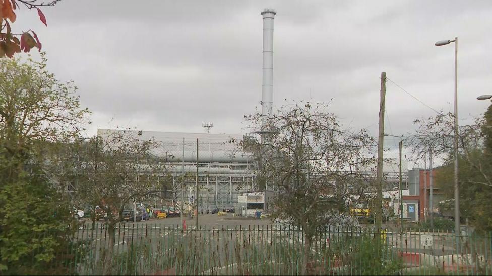 A tall chimney stands above the waste incinerator plant at Polmadie. There is a cloudy sky and trees in the foreground.