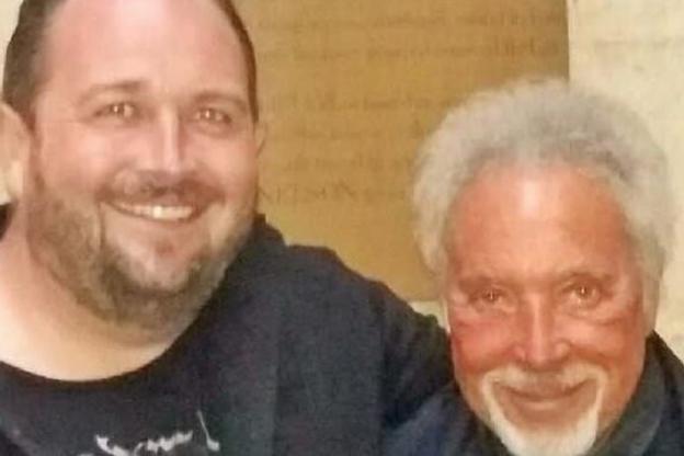 Aaaron Howlett (left) and Tom Jones (right). Aaron has brown hair and a brown stubble beard and has his arm around Sir Tom. Tom has silver hair, beard and blue eyes and they both smile at the camera.