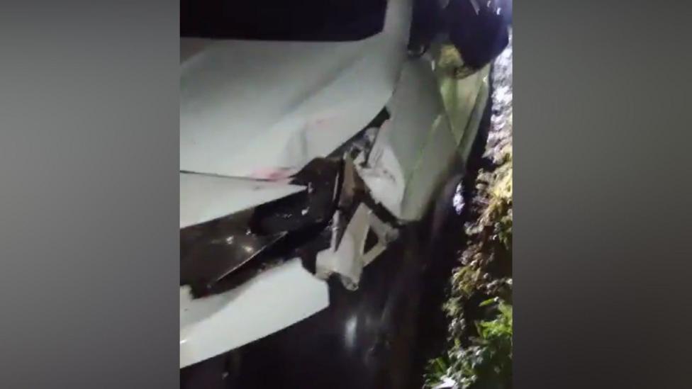 A grainy image of the front of a white van. It is dark and the van has come to a stop beside a grassy area. The front left light of the van is smashed and the body work has also been damaged around the light.