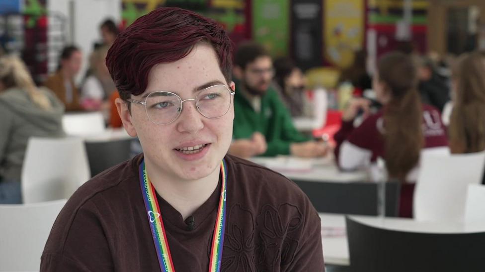 A female university student, with short dyed red hair and wearing a pair of glasses. She is sitting at a canteen on campus.
