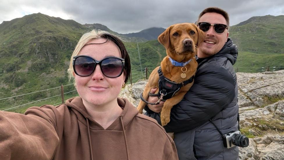 A selfie photograph of Lauren Evans (left) and her boyfriend and dog (right) in the green mountains in north Wales. Lauren has blonde and black hair and smiles at the camera wearing black wide sunglasses. She wears a brown hoodie while her boyfriend wears a black and grey puffer coat and has a camera strapped across his body while he holds their dog in his arms. He has brown short hair and wears black sunglasses.