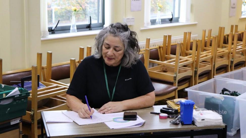Woman with grey hair in a black t-shirt with a green lanyard. She is at a table writing on paper. There is a mini blue bin at her desk with her yellow bag, some napkins and some Vaseline. behind her is stacked up chairs.