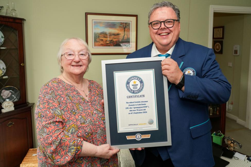 Two people stand in a living room holding a framed Guinness World Records certificate. One person wears a patterned top and the other a blue Guinness World Records blazer. The certificate recognizes the "oldest Fortnite streamer (female)" record, awarded to a 78-year-old streamer as of 1 September 2026. A display cabinet and framed artwork are visible in the background.
