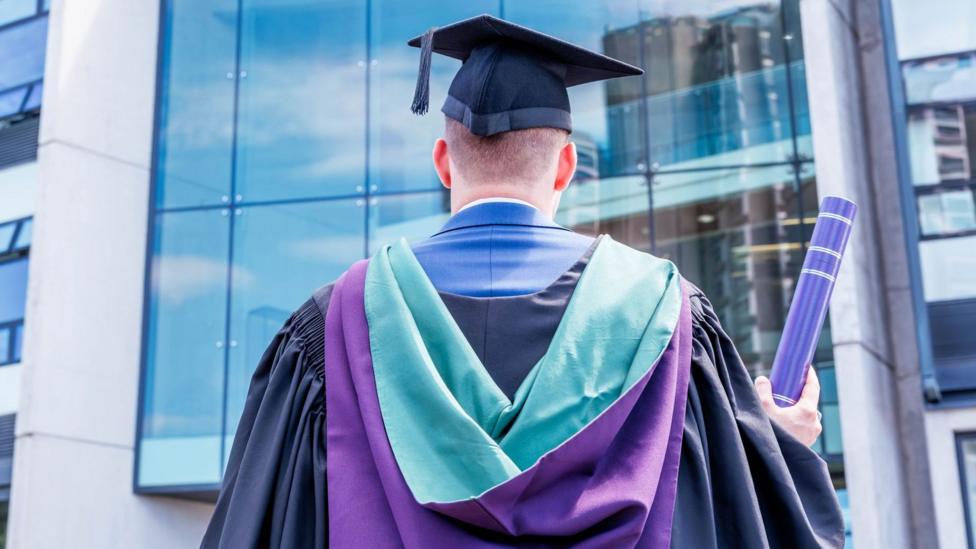 A man with his back to the camera. he is standing in front of a large building. he is dressed in green and purple graduation ropes and is holding a purple tube containing his diploma.
