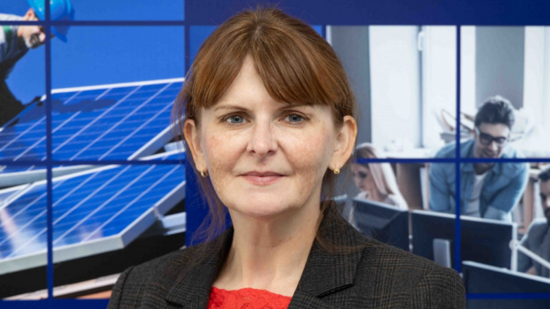 Caoimhe Archibald is smiling at at the camera. She is wearing a grey blazer and red blouse. She has ginger hair, which is tied back. 