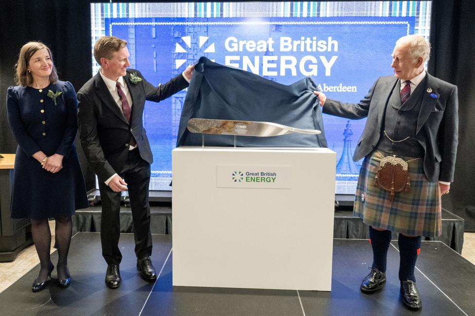 The King, dressed in a kilt, unveils a metal plaque with a man and a woman standing in the left of the photo