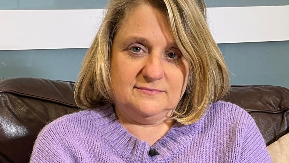 A woman is pictured looking at the camera while wearing a purple jumper, sat on a sofa.