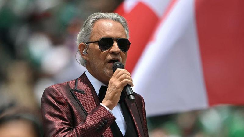 The classical singer Andrea Bocelli is pictures wearing a red patterned suit with a tuxedo. He has grey hair slicked back, and is holding a microphone in his hand.