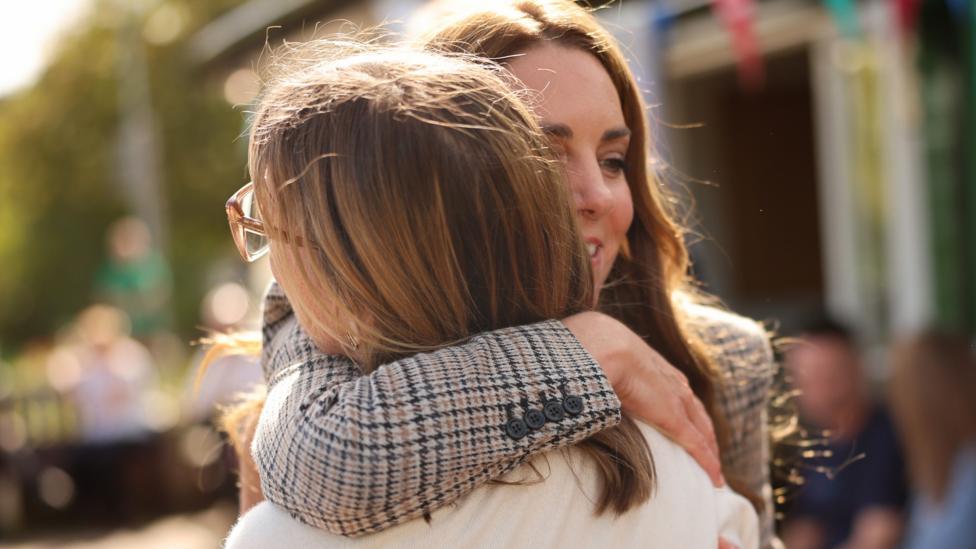 Catherine, Princess of Wales, facing the camera but partially obscured, puts her right arm around a woman's shoulders to pull her into a hug. She has long brown hair and a grey Tweed-style jacket. The woman has her back to the camera. She has shoulder-length brown hair, glasses and a cream top.