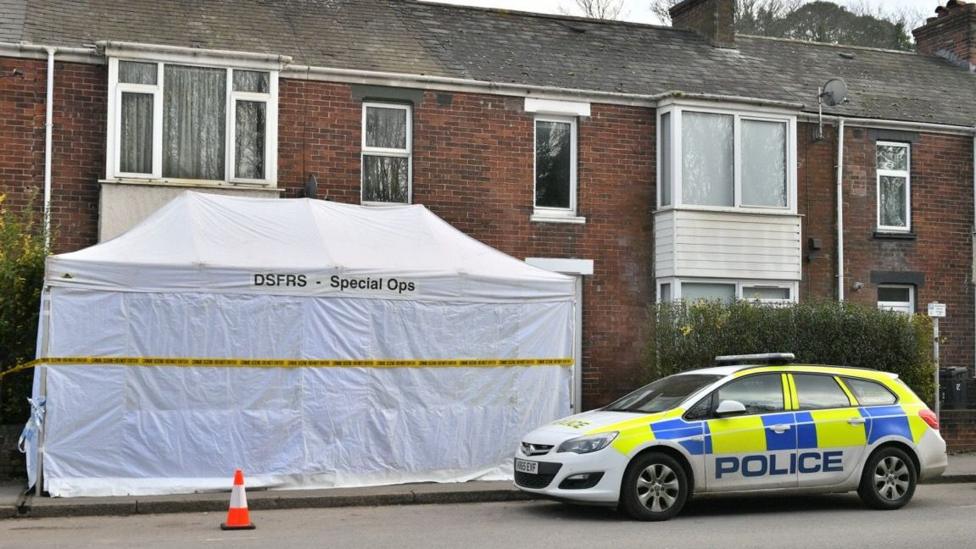 Image shows row of red brick terrace houses with white UPVC windows. There is a large white tent in front of one of the houses and a blue and yellow police car is parked to the right of it. There is also a traffic cone in front of the tent.