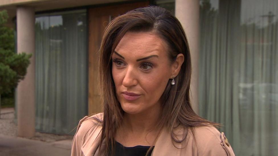 Claire has dark brown hair and is wearing a a neutral coloured jacket.