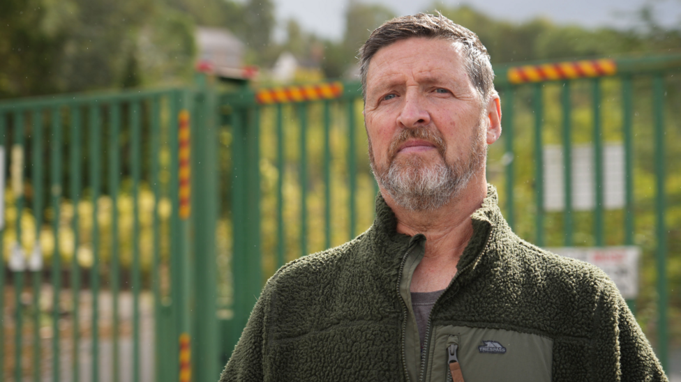 Michael Phillips is a white man with brown hair and a brown and white beard wearing a green jacket in front of a green fence and woodland
