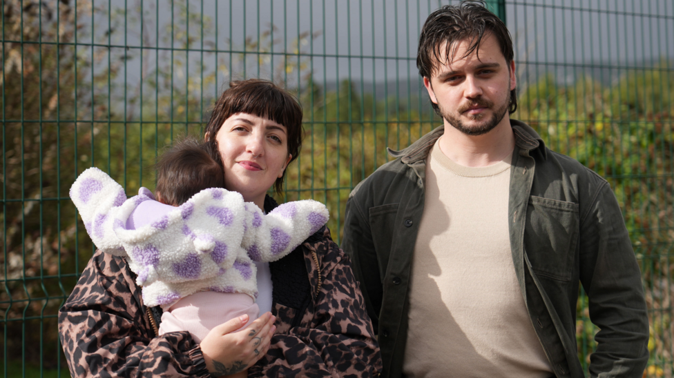 Nel Byers, her fiancé Otis Proctor and their three month old daughter Wren in front of a green fence
