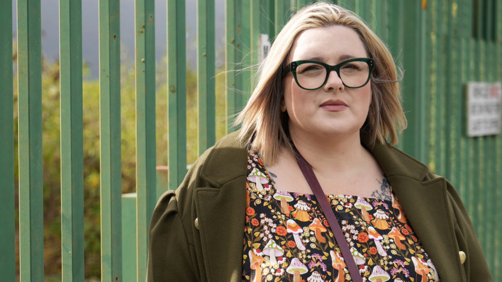 Community councillor Katie Witherden is a white woman, with blonde hair and black glasses standing in front of a green fence wearing a green coat and a pattern dress