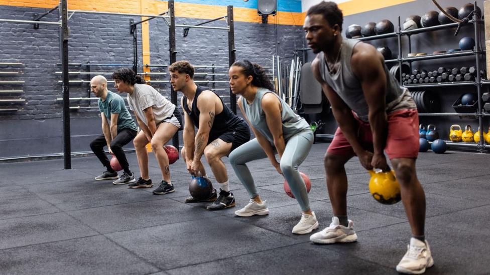 People in a gym are lined up using kettle bells, swinging them between their legs with feet apart - behind them are shelves of weights including dumb bells and bar bells. The walls are painted grey. The floor mats are also grey.