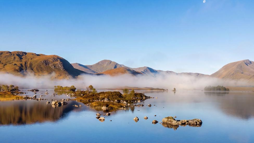 A tranquil mountain lake reflects rolling hills and scattered rocky islets beneath a clear blue sky. A layer of low mist drifts across the water, adding a serene and atmospheric touch to the landscape.