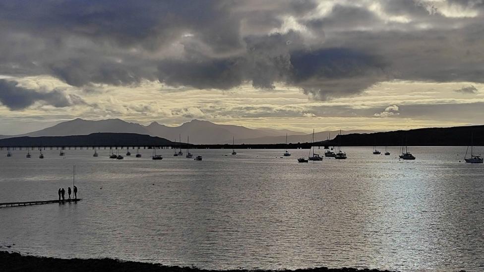 Dark, dramatic clouds hang over a calm bay where numerous sailboats lie anchored on silvery water, with distant mountains forming a layered horizon. A long bridge stretches across the background, while a small group stands at the end of a narrow pier in the foreground. Soft evening light breaks through the cloud cover, reflecting across the water and creating a moody yet tranquil coastal scene.