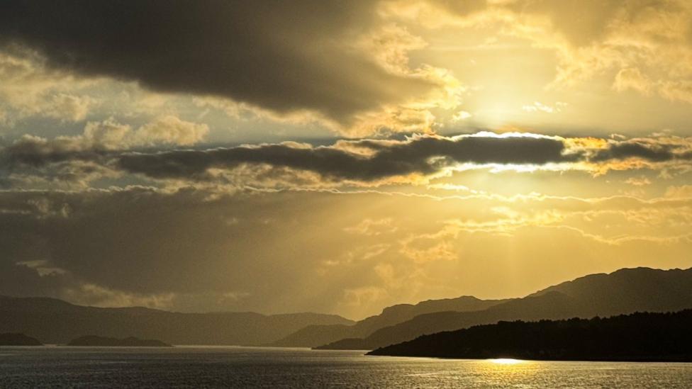 Golden sunlight streams through layered clouds, casting radiant beams across a tranquil expanse of water and silhouetted hills. The warm glow reflecting on the bay contrasts beautifully with the dark cloud formations, creating a dramatic and atmospheric landscape.