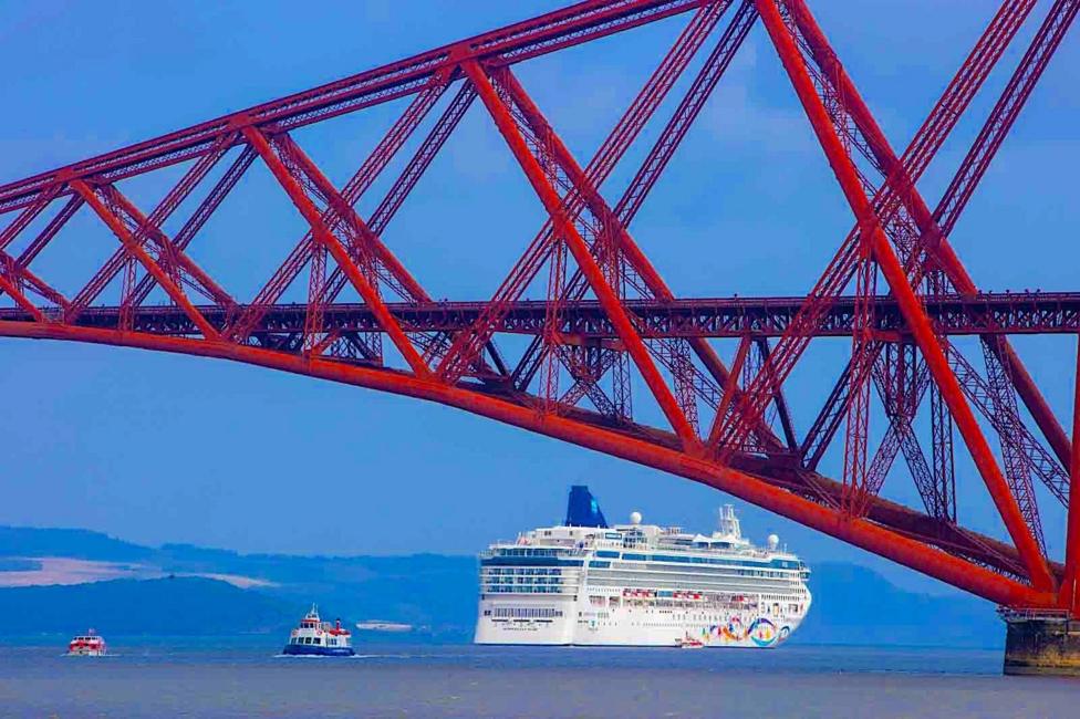 A striking red steel cantilever bridge spans across calm waters as a large white cruise ship sails beneath it. The vivid colours of the bridge contrast with the blue sky and sea, creating a dramatic coastal scene.