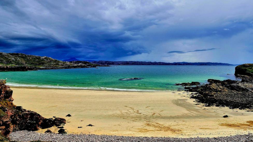 A pristine sandy beach curves around a turquoise bay, framed by rugged rocky outcrops and crystal-clear coastal waters. Dramatic storm clouds sweep across the sky, creating a striking contrast between the darkening weather and the bright, tranquil shoreline.
