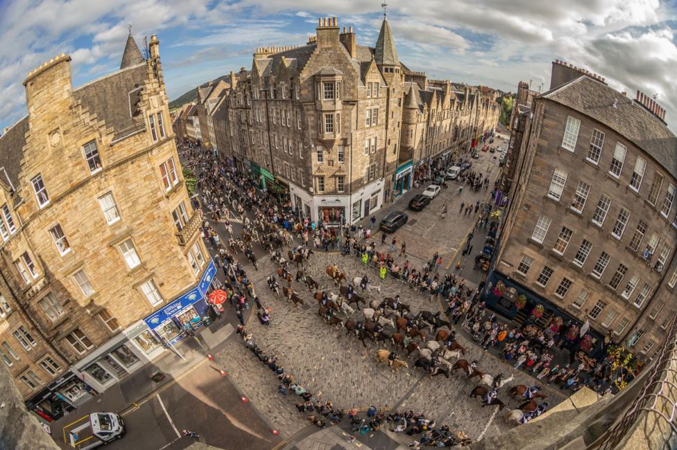 A dramatic high-angle view captures the Ride of the Marches procession winding through Edinburgh’s historic Old Town, with riders on horseback crossing a crowded cobbled junction between grand stone buildings. Spectators line the streets and gather at corners to watch the traditional event, while the curved perspective emphasises the scale, architecture and spectacle of the city-centre parade.