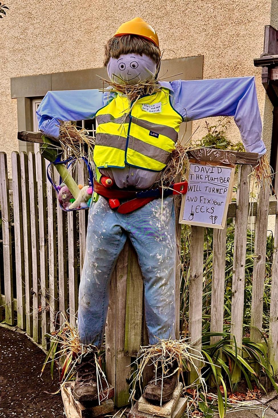 A whimsical scarecrow dressed as a plumber stands by a garden fence, wearing a high-visibility vest, hard hat and tool belt stuffed with oversized tools. A humorous sign reads, “David the Plumber will turnip and fix your leeks,” using vegetable-themed wordplay to create a charming and creative display.