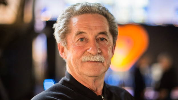 Philip King has short white hair and a moustache. He is wearing a black top and is standing side on. The background is blurred.