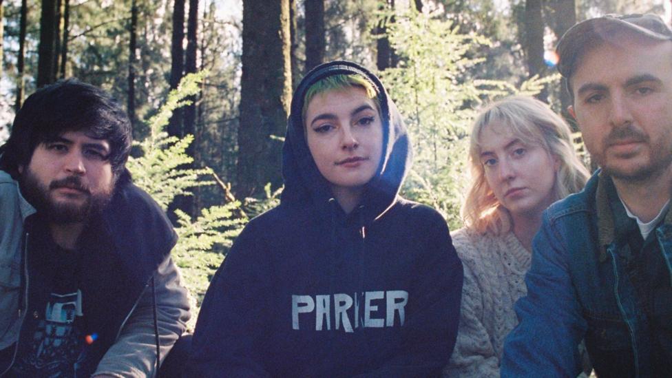 A number of individuals are pictured in the woods. Some are wearing coats and hoodies and a number of trees are visible in the background
