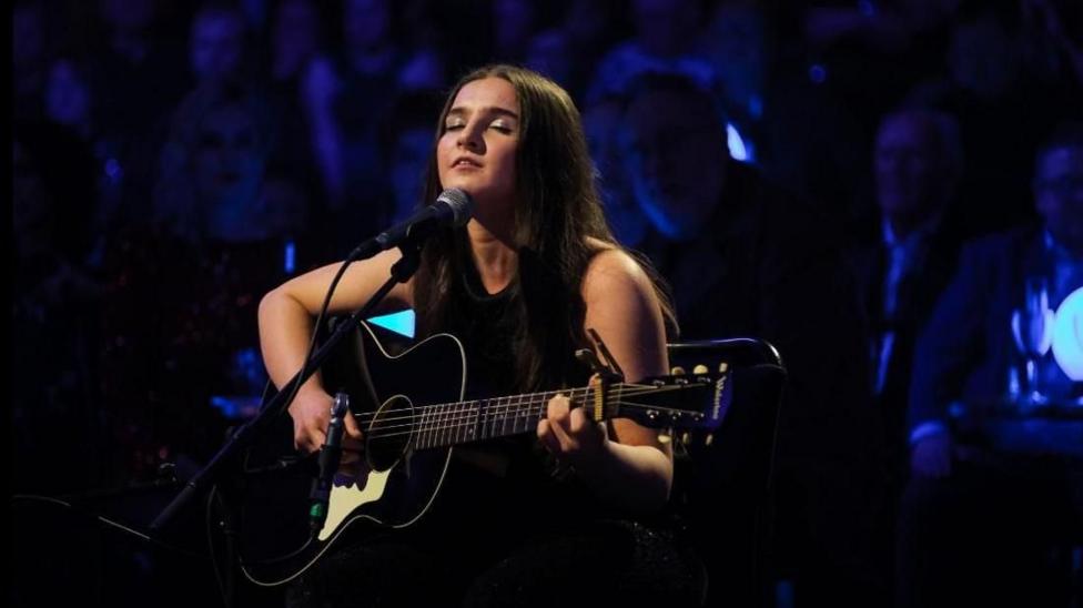 Muireann Bradley has long brown hair and is wearing a black top and holding a large guitar while performing on Jools' Annual Hootenanny