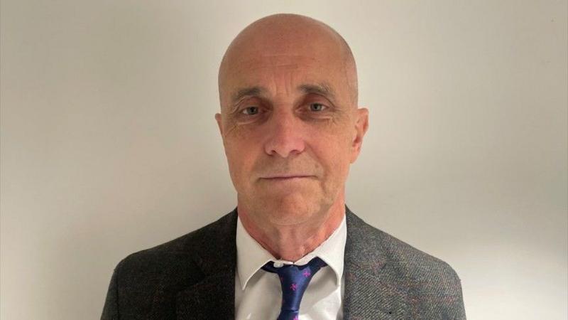 Steve Spoerry, a man with shaven head, with grey jacket, white shirt and blue tie.