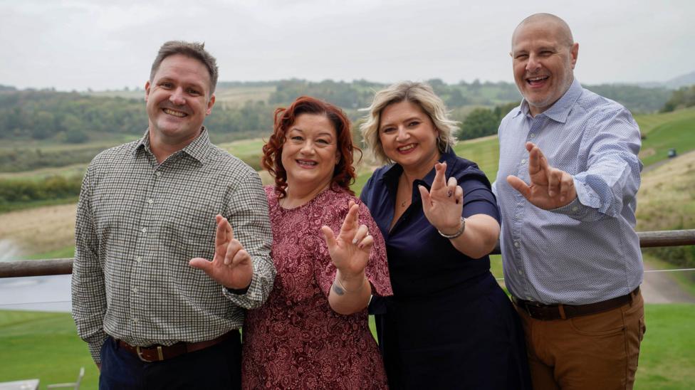 Gareth, Amanda Louise and Matthew (left to right) standing side by side and all crossing their fingers mimicking the National Lottery logo, with arms stretched out towards the camera and big smiles. 