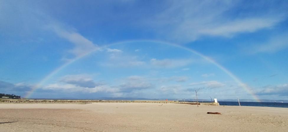 Wide sandy beach beneath a bright rainbow arching across a blue, cloud-filled sky.
