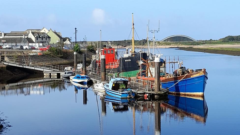 Fishing boats moored in a calm harbour, reflected in still water beneath a clear blue sky.