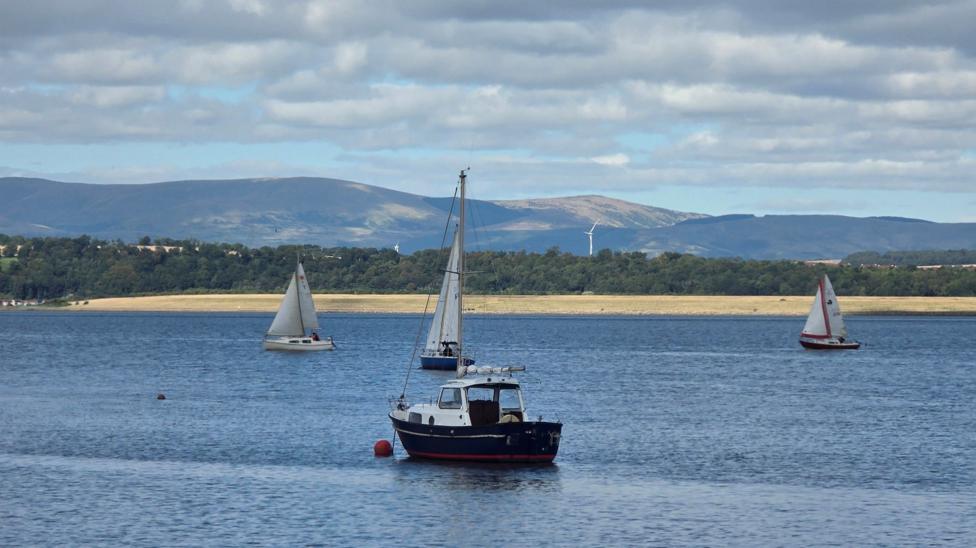 Small boats and sailboats on calm water with hills and scattered clouds in the background.