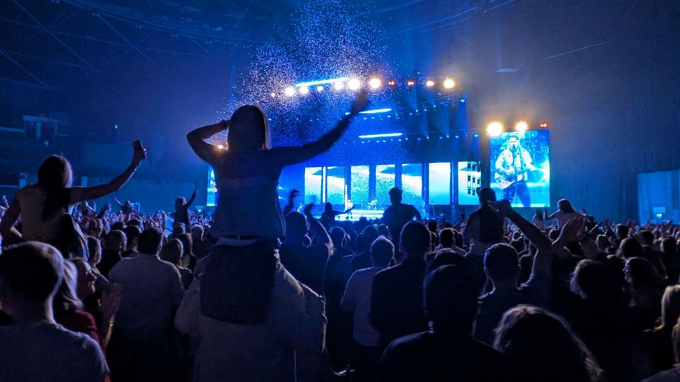Crowd at an indoor concert, illuminated by blue stage lights and large video screens.