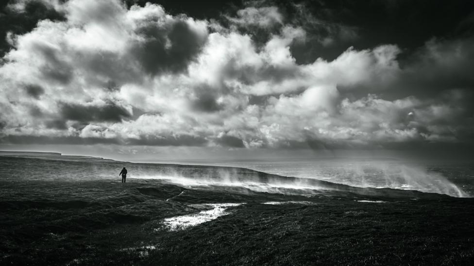 Lone figure on a windswept coastline beneath dramatic clouds in a black-and-white landscape.