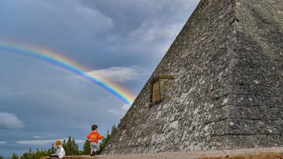 Two children sit and play beside a large stone fortification as a bright rainbow arcs across a cloudy sky.