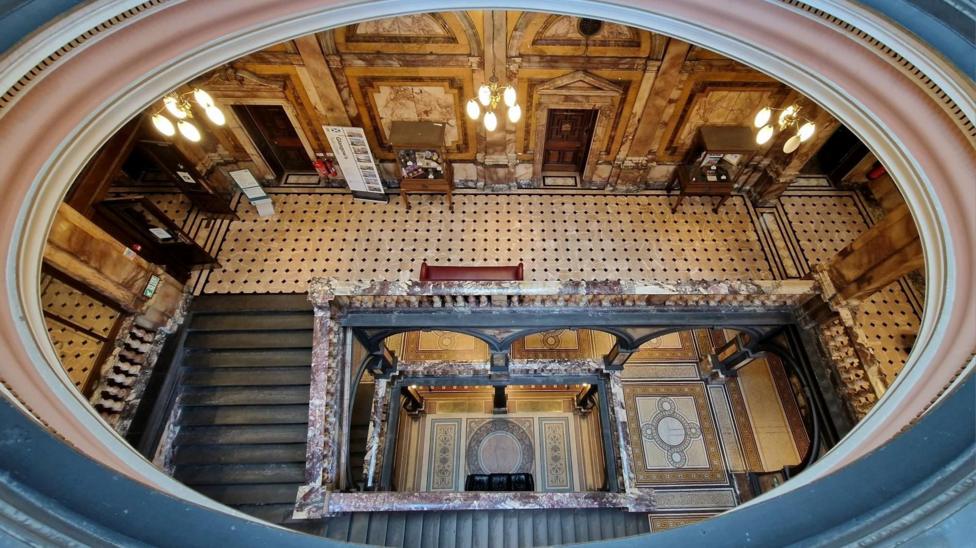 View from above of an ornate historic building interior, showing a grand staircase, patterned tiled floor and decorative marble details.