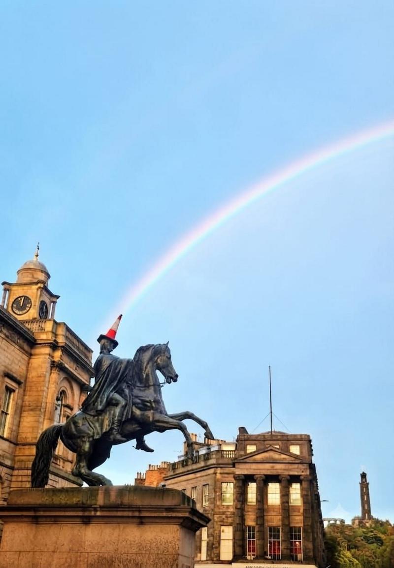 Equestrian statue wearing a traffic cone beneath a rainbow in a city square.