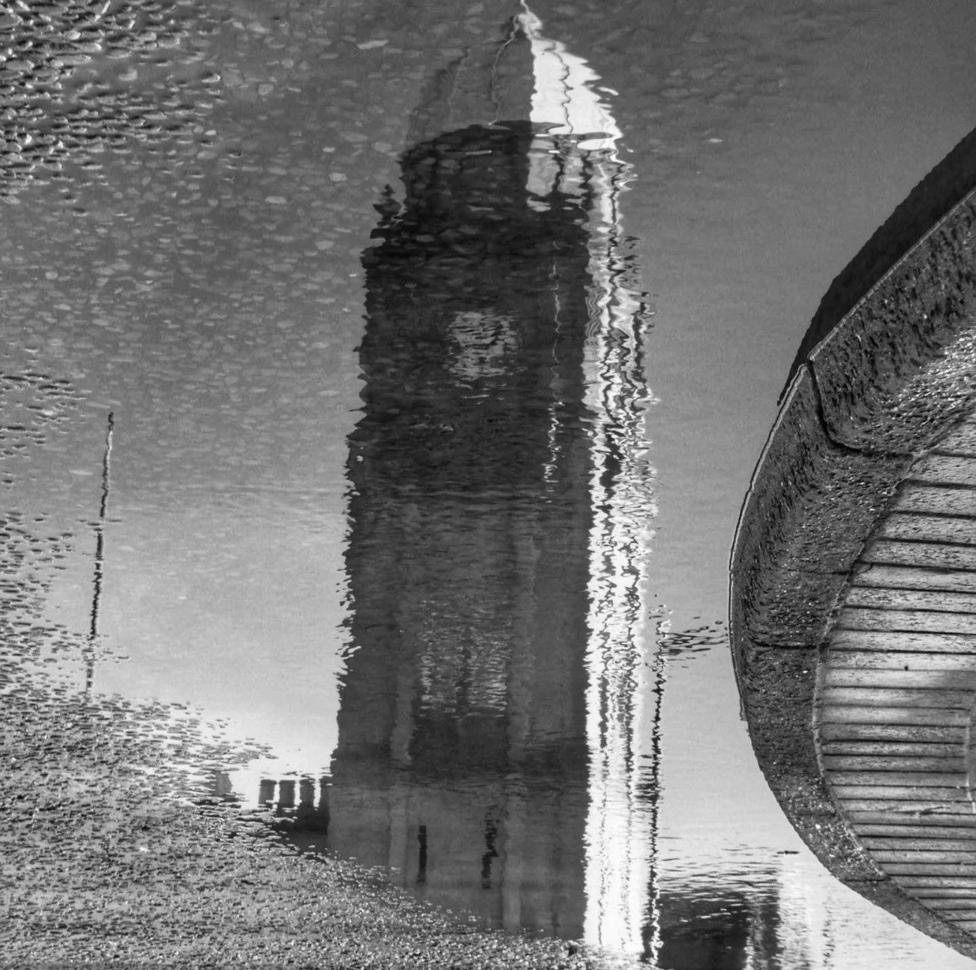 Black-and-white reflection of a lighthouse mirrored in calm water beside a curved sea wall.
