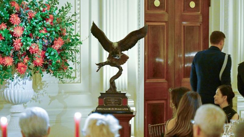A statue of a bald eagle is mounted on a podium in the East Room of the White House with seated guests looking on. The eagle has its wings outstretched as if in flight.