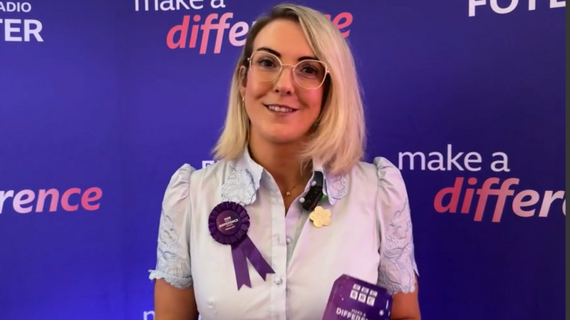 A woman with blonde hair, she has glasses and is wearing a purple badge